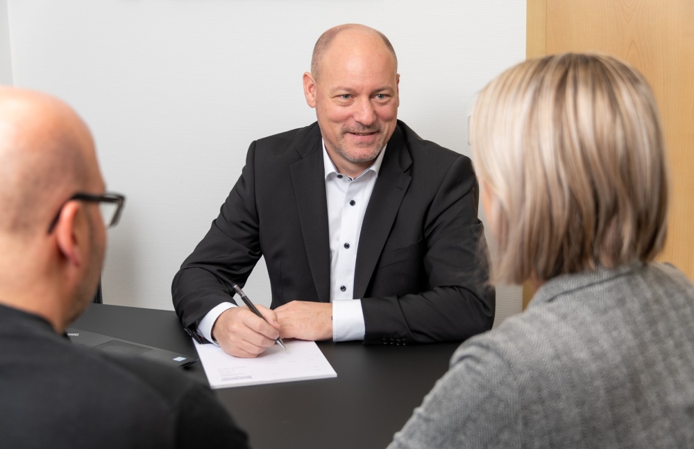 Mitarbeiter der Steuerkanzlei sitzt mit zwei Mandanten an einem Tisch und führt ein Beratungsgespräch, mit Stift und Notizblock in der Hand.