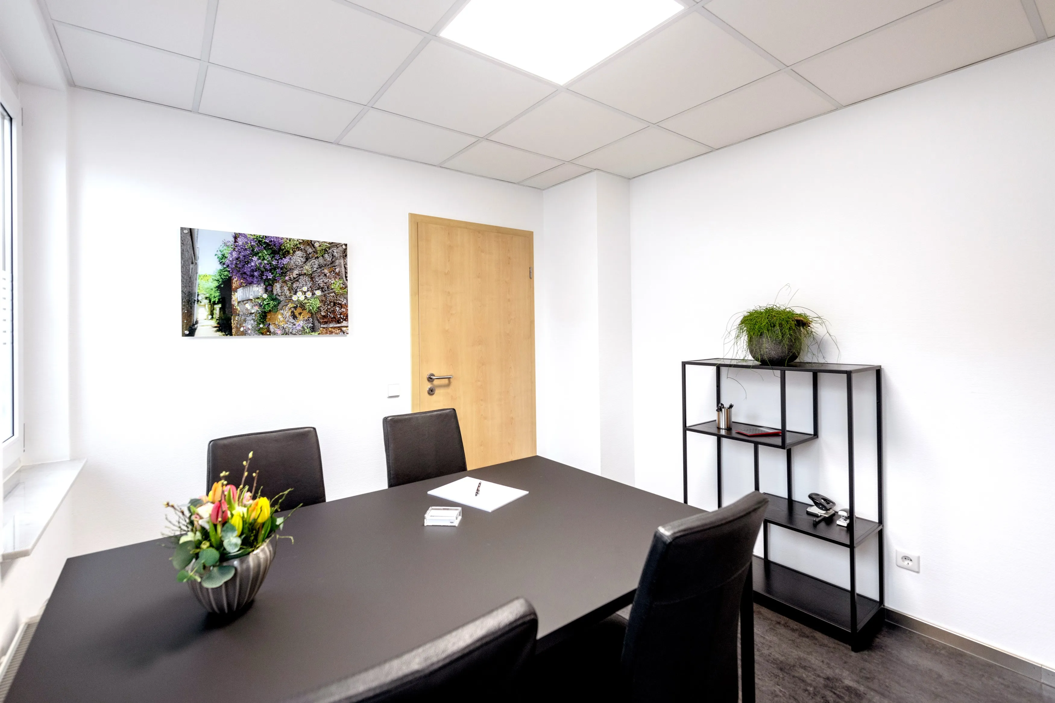 Moderner Besprechungsraum der Steuerkanzlei mit schwarzem Tisch und Stühlen, Regal mit Pflanze und Wandbild.