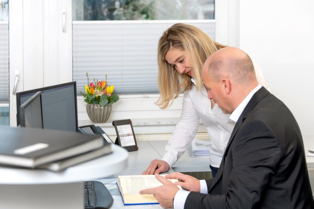 Zwei Mitarbeitende der Steuerkanzlei arbeiten gemeinsam am Schreibtisch, schauen in ein aufgeschlagenes Buch und besprechen Inhalte.