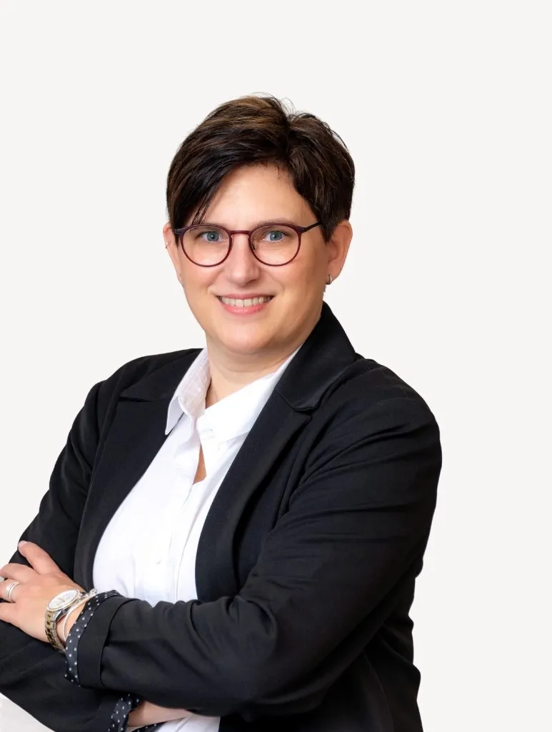 Porträtfoto einer Mitarbeiterin der Steuerkanzlei im schwarzen Blazer und weißem Hemd, mit kurzer dunkler Frisur und Brille, freundlich lächelnd.